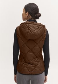 Gilet matelassé brun avec capuche, présentant des motifs en diamant et une silhouette ajustée, porté par-dessus un haut à manches longues noir.