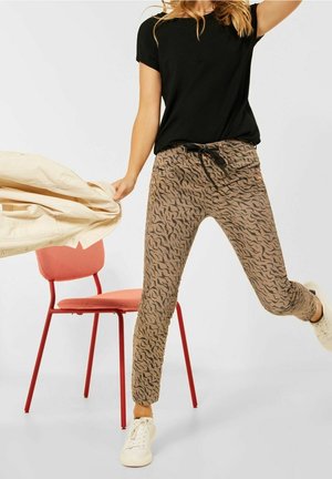 Vrouw die beige broek met patroon, zwart T-shirt en witte sneakers draagt, een beige jas vasthoudt, staand bij een rode stoel.