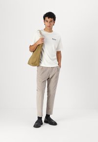 Béžová plátěná taška, bílý tričko s krátkým rukávem s logem, světle šedé kalhoty a černé boty. Minimalistický design, uvolněný střih.