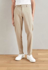 Pantalons en lin beige avec une coupe droite, dotés d'une fermeture à un bouton et de plis à l'avant. Associés à des baskets blanches.