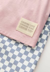 Top en coton rose avec une étiquette texturée indiquant "CHOISIS L'AMOUR, CHOISIS LA BIENVEILLANCE, CHOISIS TOI," superposé sur un motif à carreaux bleu et blanc.