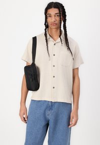 Short-sleeve, beige textured shirt with front pocket, paired with light blue denim pants. A small black crossbody bag is worn on the shoulder.