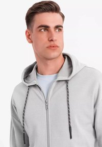 Haut de hoodie zippé gris clair avec une capuche doublée, un tissu texturé, un cordon de serrage contrastant, et un t-shirt bleu clair uni en dessous.
