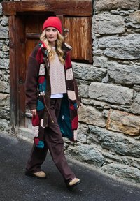 Žena kráčajúca vonku, oblečená v pestrofarebnom patchworkovom kabáte, červenej pletenej čiapke, béžovom svetri, hnedých nohaviciach a svetlohnedých topánkach pri kamennej stene a drevených dverách.
