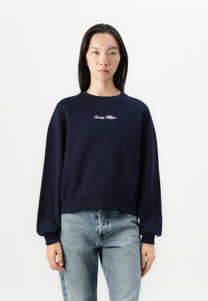 Navy blue cropped sweatshirt with ribbed cuffs and hem, featuring white embroidered branding on the chest. Worn with light denim jeans.