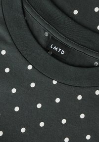 Mørkegråt stof med hvide polkaprikker og et nærbillede af en halsudskæring, der viser en sort tøjlabel med hvid "LMTD" tekst.