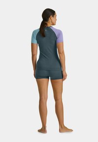 Rash guard in teal and lavender, featuring short sleeves, fitted design, and a smooth texture. Shorts in matching color complete the set.