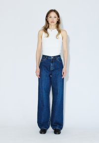 Ung kvinde med langt hår iført en hvid ærmeløs top, blå jeans med vide ben og sorte sko, står foran en hvid baggrund.
