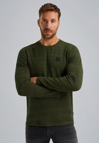 Dunkelgrünes Langarmshirt ausTextilgewebe, runder Ausschnitt und kleiner schwarzer Logopatch auf der Vorderseite. Getragen mit dunkelgrauen Jeans.