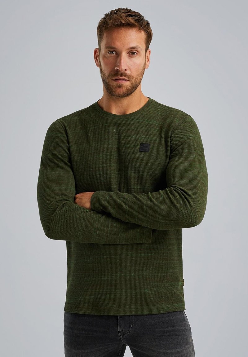 Dunkelgrünes Langarmshirt ausTextilgewebe, runder Ausschnitt und kleiner schwarzer Logopatch auf der Vorderseite. Getragen mit dunkelgrauen Jeans.