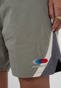 Personne portant un short gris et blanc avec un logo rouge, bleu et gris et le texte « PROHIBITED » brodé près de l'ourlet.