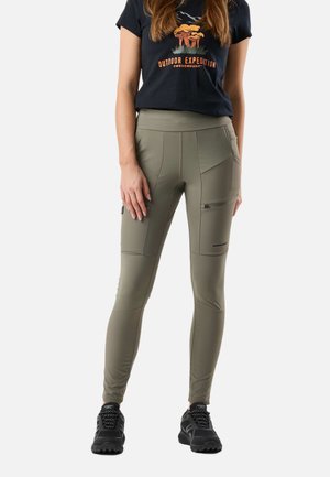 Kvinde iført olivengrønne tætsiddende cargobukser med lynlåslomme og en sort t-shirt med orange udendørsgrafik, stående i sorte sneakers.