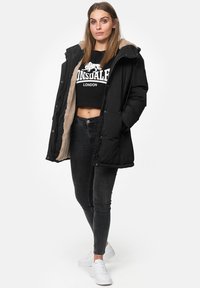 Zwarte gewatteerde jas met een Pluche beige interieur, voorzien van een ritssluiting, een kap en drukknoopsluitingen. Gekozen over een zwarte cropped sweatshirt en jeans.