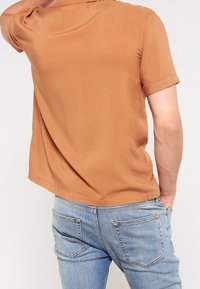 Kortärmad, brun, lätt skjorta med rak skärning. Matcher med ljusblå denimjeans som har bakfickor och subtil sömnad.