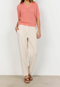 Vaaleanpunainen neulottu V-kauluspaidat lyhyillä hihoilla, yhdistettynä löysiin kermaan värisiin housuihin ja ruskeisiin remmissandaaleihin. Tasainen tausta.