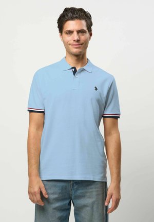 Man met kort donker haar, gekleed in een lichtblauw poloshirt met gestreepte mouwranden en een blauwe spijkerbroek, staand tegen een effen achtergrond.