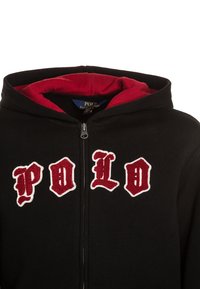 Černá mikina na zip s červenou podšívkou. Na přední straně se nachází bílý a červený nápis "P O L O". Vyrobena z měkké, texturované látky.