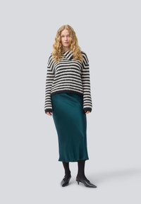 Kvinne står, iført en svart og hvitstripet genser, en teal satin midi-skjørt, svarte strømpebukser og svarte spisse kitten heels.