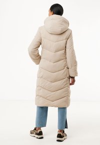 Beige lange pufferjas met chevronstiksel, hoge kraag en capuchon. Gecombineerd met lichtblauwe opgerolde jeans en bruine sneakers.