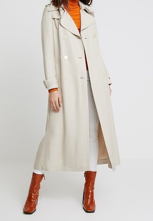 Femme portant un long trench-coat beige, un col roulé orange, un pantalon blanc et des bottines marron brillantes, debout devant un fond uni.