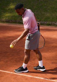 Jugador de tenis con polo rosa y pantalones cortos grises, sosteniendo una raqueta, preparándose para servir una pelota de tenis amarilla en una cancha de arcilla.