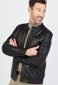 Man in een zwart leren jasje over een beige coltrui, met gekruiste armen, kijkend naar rechts met een lichte glimlach.