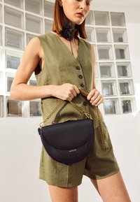 Frau in olivgrünem ärmellosem Overall hält eine schwarze Lederhandtasche mit goldener Kettenriemen vor einem Hintergrund aus Glasbausteinwand.