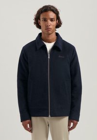 Donker marineblauwe fleecejas met een kraag, voorkant met rits en zijzakken. Voorzien van een subtiele logogegevens op de borst.