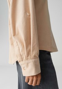 Beige corduroy long-sleeve shirt with buttoned cuffs, featuring vertical ridges and a relaxed fit, paired with dark denim pants.