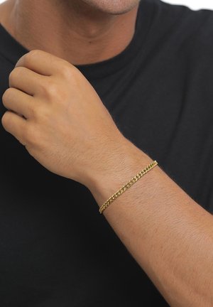Männliches Handgelenk trägt ein dünnes Goldkettenarmband mit schwarzem Hemdhintergrund, Hand teilweise zur Faust geballt in der Nähe des Gesichts.