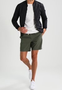 Giacca bomber nera, t-shirt grigia chiara, pantaloni corti verde oliva e sneakers bianche. Outfit casual con textures morbide e design semplice.