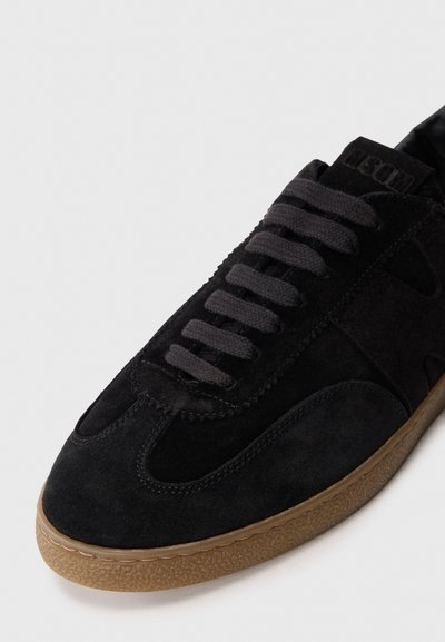 Black suede sneaker featuring a rounded toe, textured gum sole, and flat laces. Notable accents include side panel cutouts and stitching details.