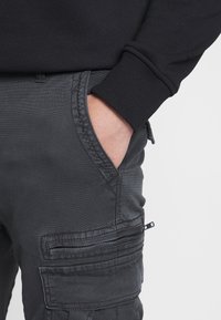 Tmavě šedé cargo kalhoty s texturovaným povrchem, které mají boční kapsy a kapsu na zip na stehně. Kombinované s černou mikinou s manžetami.