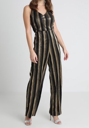 Kobieta ubrana w czarny bezrękawnikowy kombinezon z pionowymi, metalicznymi złotymi paskami, w białych sandałach na wysokim obcasie, stojąca na jednolitym tle.