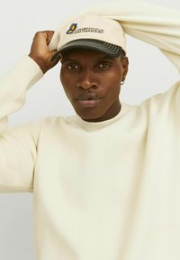 Casquette beige clair avec visière noire et logo "Originals" ; pull ivoire uni avec col en côtes, texture du tissu visible, sans motifs.