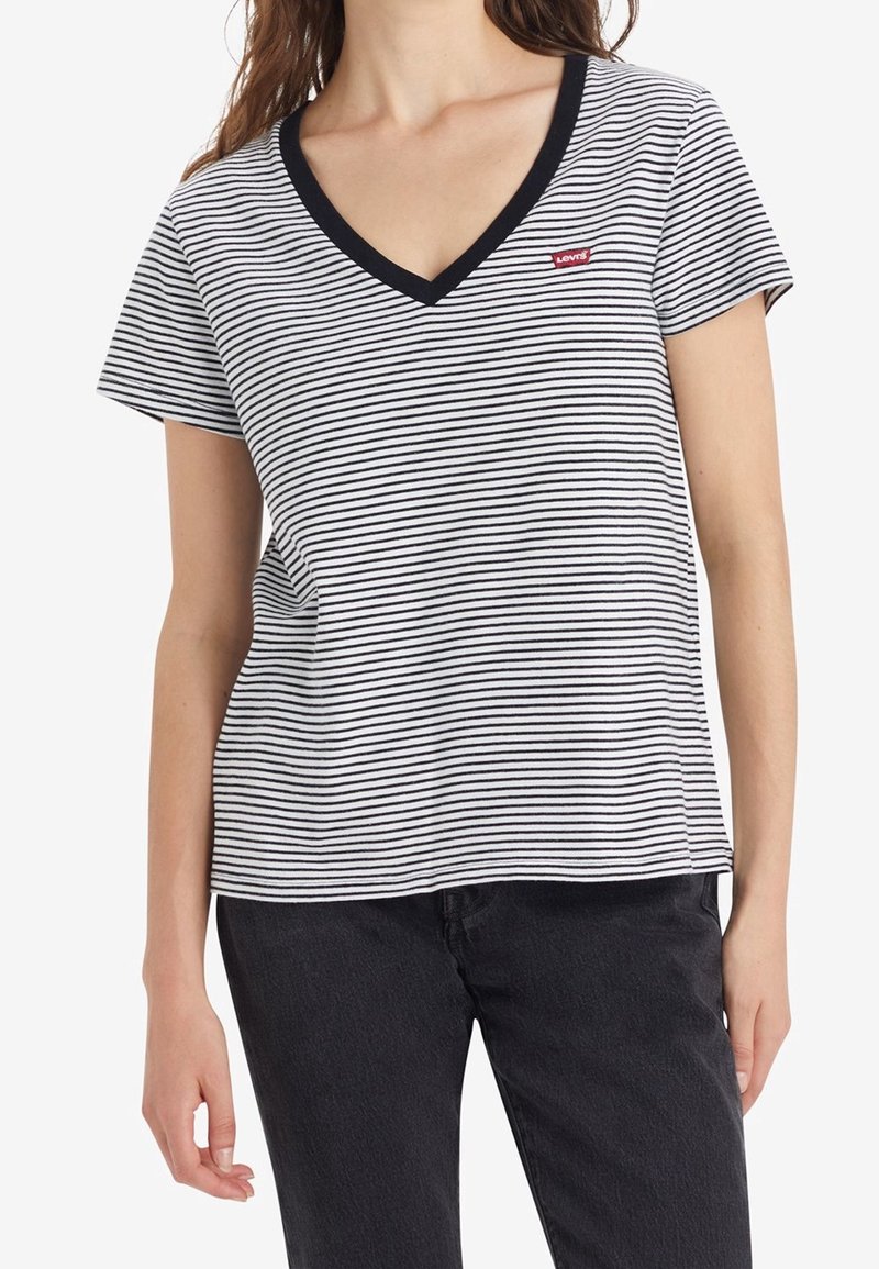Femme portant un t-shirt à rayures horizontales blanches et noires avec un col en V et un petit logo rouge Levi's sur la poitrine gauche, associé à un pantalon noir.