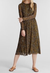 Långärmad, brun blommig klänning med utsvängd kjol. Har mönstrad textur med orange, blå och vita detaljer, kombinerat med bruna sandaler.