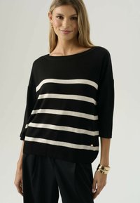 Donna sorridente che indossa un maglione nero con strisce orizzontali bianche, pantaloni neri e un bracciale dorato al polso sinistro, su uno sfondo neutro.