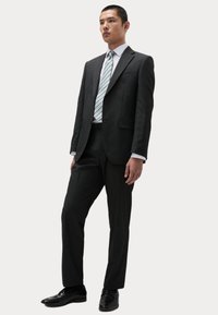 Traje ajustado de color gris oscuro con solapas muestradas, combinado con una camisa blanca y una corbata a rayas azul claro. Unos zapatos de cuero negros completan el conjunto.