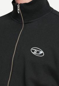 Giacca nera con zip frontale, colletto alto, texture a coste e cerniera argentata. Presenta un logo ovale metallico sul petto.