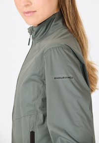 Lichte, olijfgroene jas met een hoge kraag. Beschikt over een ritsluiting, een gladde textuur en het "ENDURANCE" logo op de mouw.