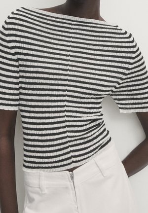 Personne portant un haut côtelé à manches courtes à rayures horizontales noires et blanches, associé à un pantalon blanc taille haute avec fermeture éclair à l’avant.