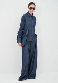 Donkerblauwe denim shirt en wijde broek, beide met zichtbare stiksels. Model houdt een marineblauwe tas vast en draagt oversized zwarte zonnebril.