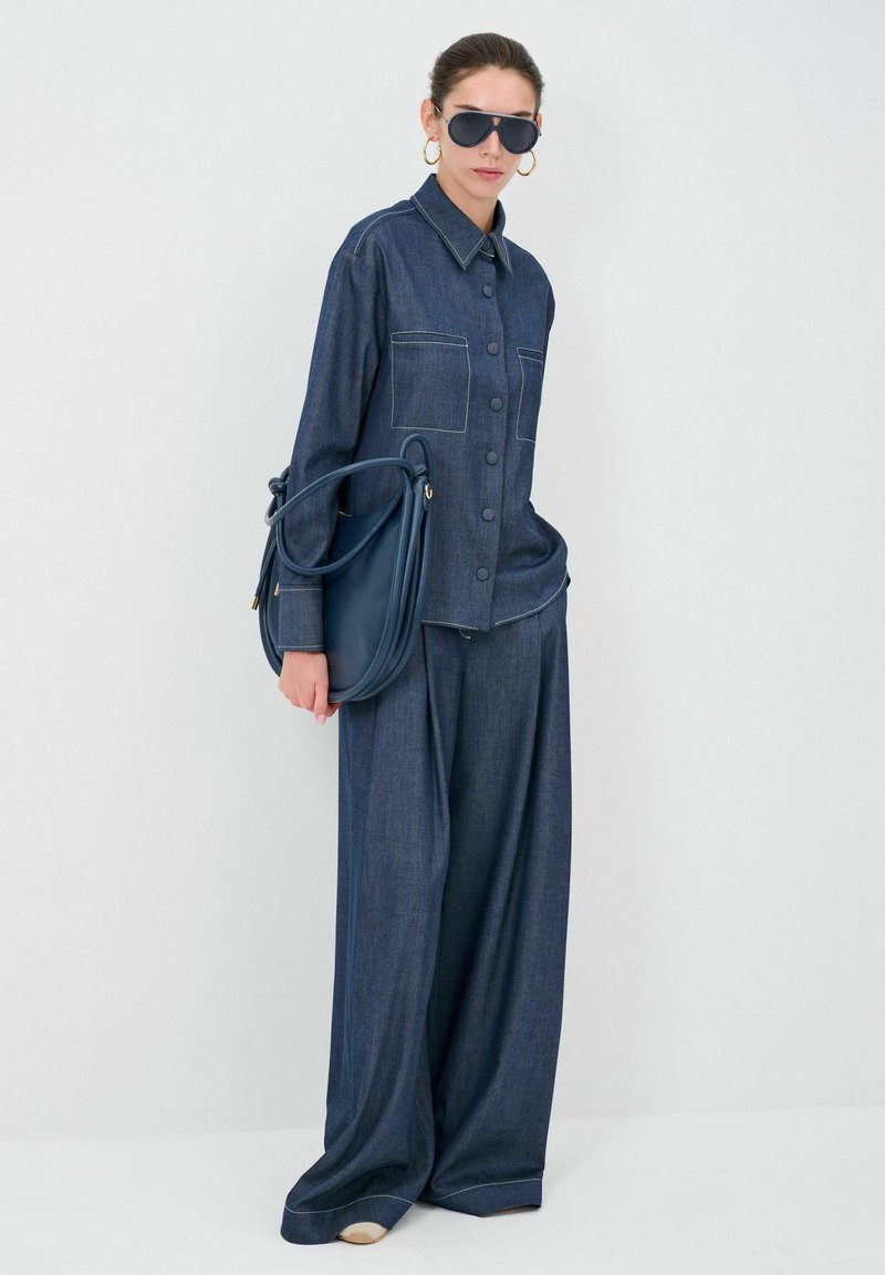 Donkerblauwe denim shirt en wijde broek, beide met zichtbare stiksels. Model houdt een marineblauwe tas vast en draagt oversized zwarte zonnebril.
