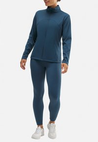 Chaqueta atlética azul con cuello alto y cierre de cremallera, combinada con leggings ajustados a juego. La tela parece suave y elástica.