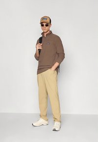 Pruun fliisist poollukkidega pullover, beežid püksid, valged vabaajajalatsid, beež nokamüts ja valged päikeseprillid, luues lõbusa vabaaja välimuse.