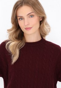 Burgunderfarbener Strickpullover mit Kabelmuster, rundem Ausschnitt und weicher Textur. Modell mit welligem Haar vor einem schlichten weißen Hintergrund.