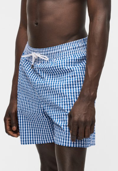 Άνδρας που φοράει μπλε και λευκά καρό μαγιό με λευκό κορδόνι, εμφανιζόμενος από τη μέση έως το μέσο του μηρού σε λευκό φόντο.