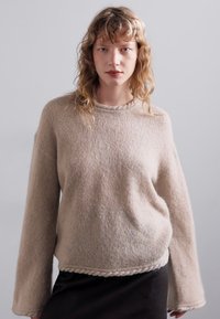 Bézs színű, oversized pulóver, texturált kötött anyagból készült, kerek nyakkal és fonott szegéllyel. Hosszú ujjak járulnak hozzá a laza szabáshoz.