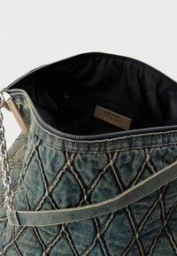 Torbica od trapera s prošivenim uzorkom, širokim otvorom, srebrnim patentnim zatvaračem i remenom od lanca. Unutrašnjost je crna s brendiranim etiketom i džepom.